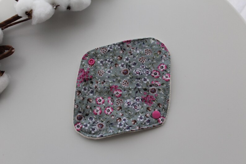 May include: A square, floral-patterned reusable pad with a gray background and pink and white flowers. The pad has two pink snaps and a white border. The pad is made of soft fabric.
