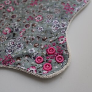 May include: Close-up of a floral patterned fabric with pink and white flowers on a muted green background. The edge is trimmed with a white border and accented with pink snaps.