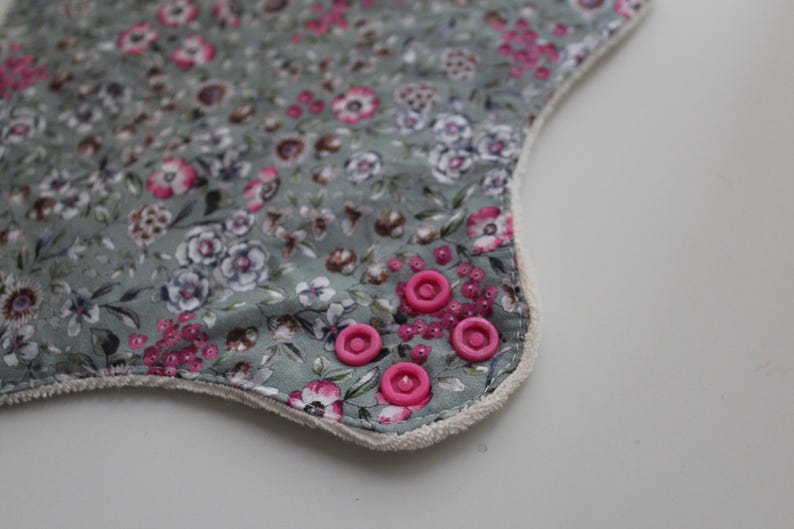 May include: Close-up of a floral patterned fabric with pink and white flowers on a muted green background. The edge is trimmed with a white border and accented with pink snaps.