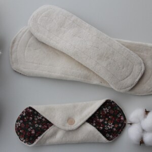 May include: An overhead shot displays two white, reusable menstrual pads and one folded pad with a floral pattern. The pads are made of soft, absorbent material. Cotton sprigs are placed on the left and right sides of the pads.