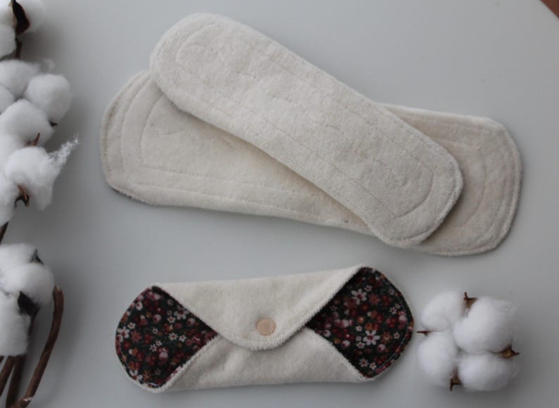 May include: An overhead shot displays two white, reusable menstrual pads and one folded pad with a floral pattern. The pads are made of soft, absorbent material. Cotton sprigs are placed on the left and right sides of the pads.
