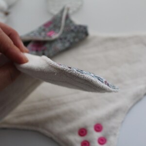 May include: A close-up of reusable cloth menstrual pads. One pad is held up, revealing a floral patterned edge. The pads are white with pink accents and pink snaps. The pads are designed for comfort and eco-friendliness.