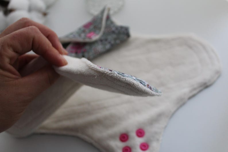 May include: A close-up of reusable cloth menstrual pads. One pad is held up, revealing a floral patterned edge. The pads are white with pink accents and pink snaps. The pads are designed for comfort and eco-friendliness.