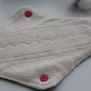 May include: A reusable, light gray cloth menstrual pad with a textured surface. The pad has a curved shape with pink snap closures and decorative stitching. The pad is designed for comfort and absorbency.