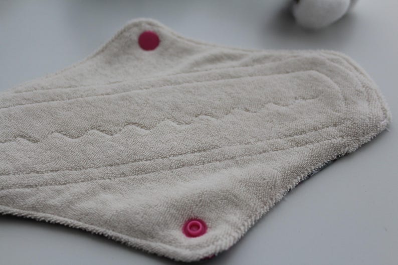 May include: A reusable, light gray cloth menstrual pad with a textured surface. The pad has a curved shape with pink snap closures and decorative stitching. The pad is designed for comfort and absorbency.