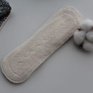 May include: A beige reusable menstrual pad with a quilted design, next to a sprig of cotton. A portion of a floral patterned pad is visible on the left.