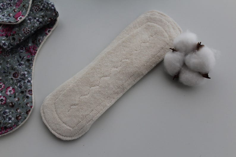 May include: A beige reusable menstrual pad with a quilted design, next to a sprig of cotton. A portion of a floral patterned pad is visible on the left.