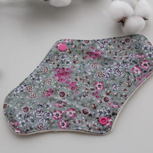 May include: Reusable cloth menstrual pads with a floral pattern in shades of gray, pink, and purple. The pads have pink snap closures and a white trim. The pads are designed for feminine hygiene.