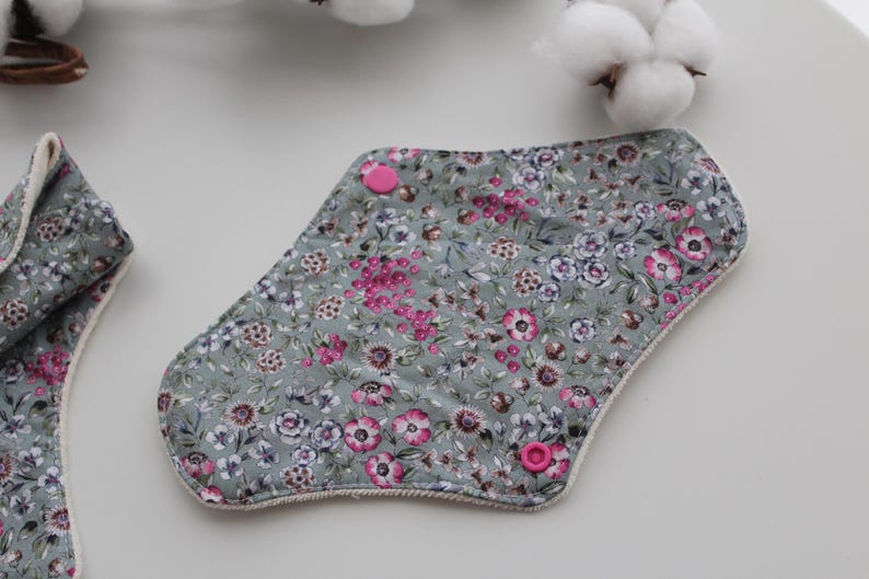 May include: Reusable cloth menstrual pads with a floral pattern in shades of gray, pink, and purple. The pads have pink snap closures and a white trim. The pads are designed for feminine hygiene.