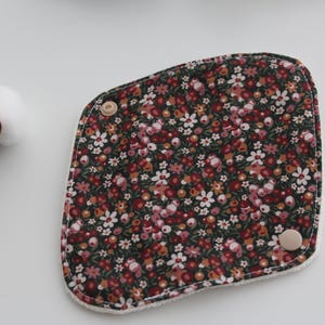 May include: A floral patterned reusable menstrual pad with a dark background and small red, pink, and white flowers. The pad has a rounded shape with two wooden buttons and a white border. Cotton buds are in the upper left corner.