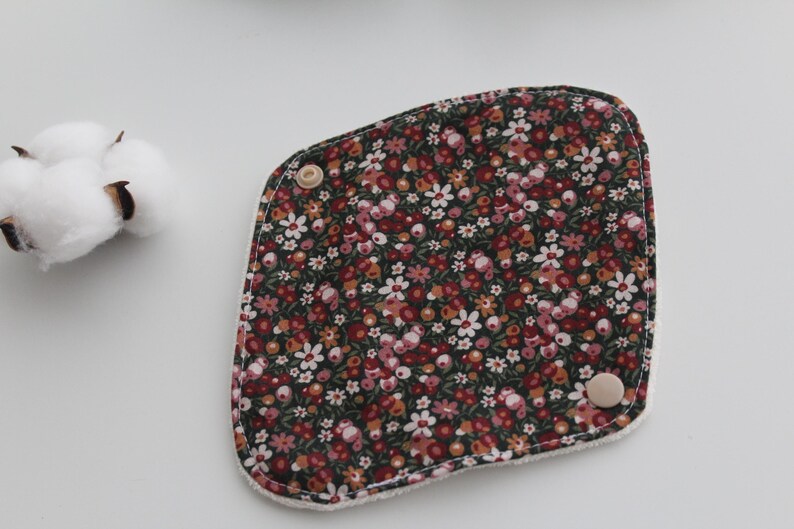 May include: A floral patterned reusable menstrual pad with a dark background and small red, pink, and white flowers. The pad has a rounded shape with two wooden buttons and a white border. Cotton buds are in the upper left corner.