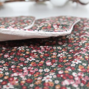 May include: Close-up of a floral patterned fabric with a white border. The fabric features a dark green background with small red, pink, and white flowers. The white border is soft and fluffy. The fabric is folded over, revealing the white interior.