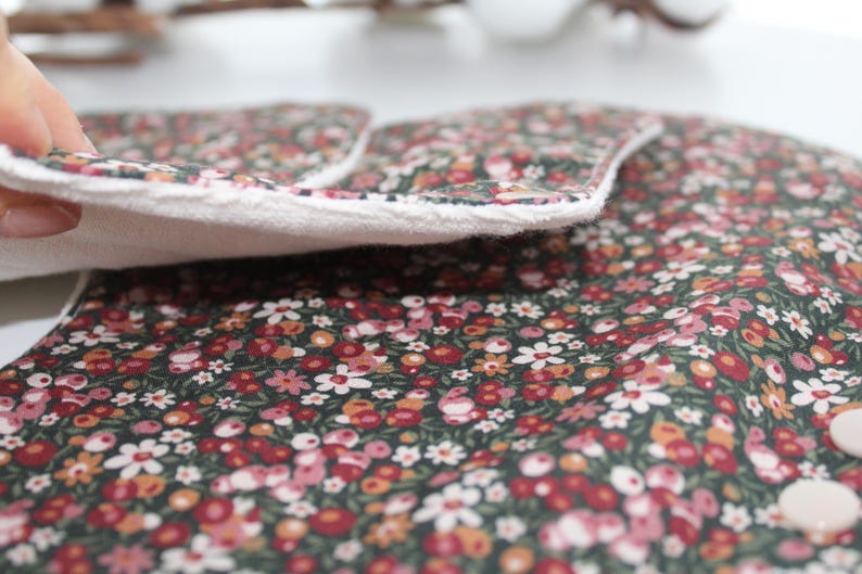 May include: Close-up of a floral patterned fabric with a white border. The fabric features a dark green background with small red, pink, and white flowers. The white border is soft and fluffy. The fabric is folded over, revealing the white interior.