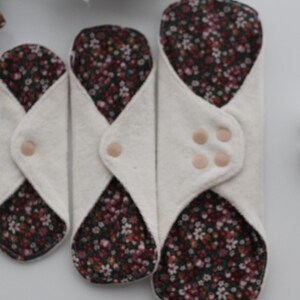May include: Three reusable cloth menstrual pads in varying sizes. The pads have a white exterior and a floral patterned interior with a snap closure. A cotton flower is visible in the background.