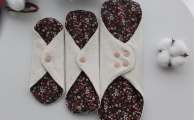 May include: Three reusable cloth menstrual pads in varying sizes. The pads have a white exterior and a floral patterned interior with a snap closure. A cotton flower is visible in the background.
