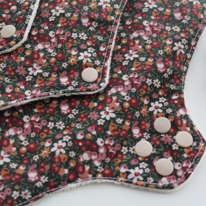 May include: Close-up of a reusable cloth menstrual pad with a floral pattern in shades of red, white, and yellow on a dark green background. The pad has white trim and several beige snaps. A cotton ball is in the background.