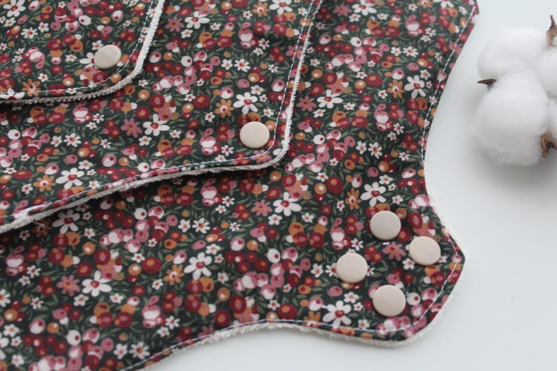 May include: Close-up of a reusable cloth menstrual pad with a floral pattern in shades of red, white, and yellow on a dark green background. The pad has white trim and several beige snaps. A cotton ball is in the background.