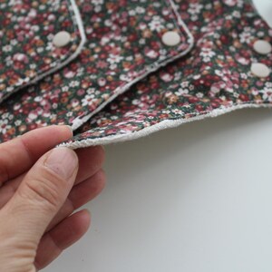 May include: Reusable cloth menstrual pads with a floral pattern in shades of burgundy, pink, and white on a dark green background. The pads have a white trim and snap closures. The image shows a close-up of the pads.
