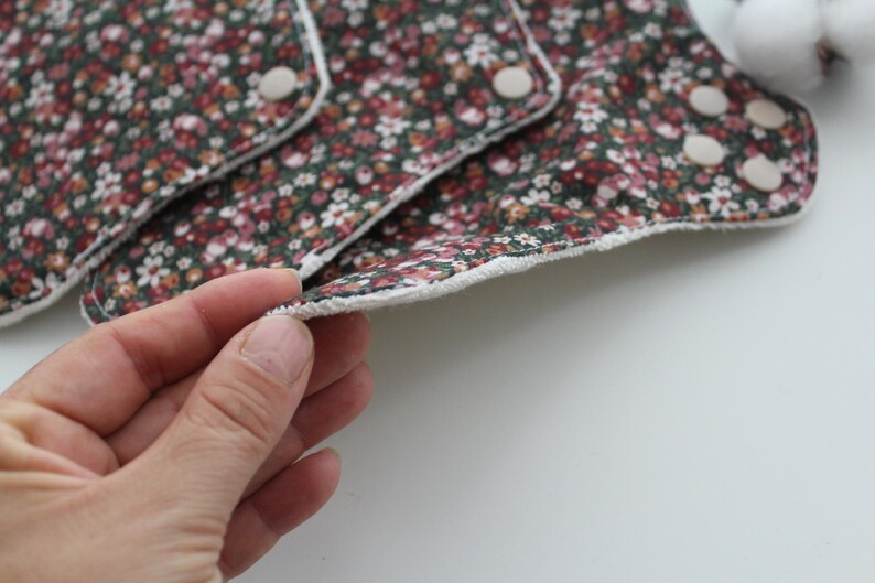 May include: Reusable cloth menstrual pads with a floral pattern in shades of burgundy, pink, and white on a dark green background. The pads have a white trim and snap closures. The image shows a close-up of the pads.