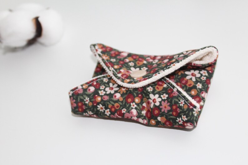 May include: A floral patterned, reusable cloth pad with a dark green background and a variety of flowers in shades of red, pink, and white. The pad is folded and secured with a wooden button.
