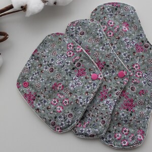 May include: Three reusable cloth menstrual pads in varying sizes, featuring a floral pattern in shades of gray, pink, and white. The pads have pink snap closures. A sprig of cotton adds a decorative touch.