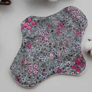 May include: Reusable cloth menstrual pads with a floral pattern in shades of gray, pink, and white. The pads have pink snap closures. A cotton ball and other pads are also visible.
