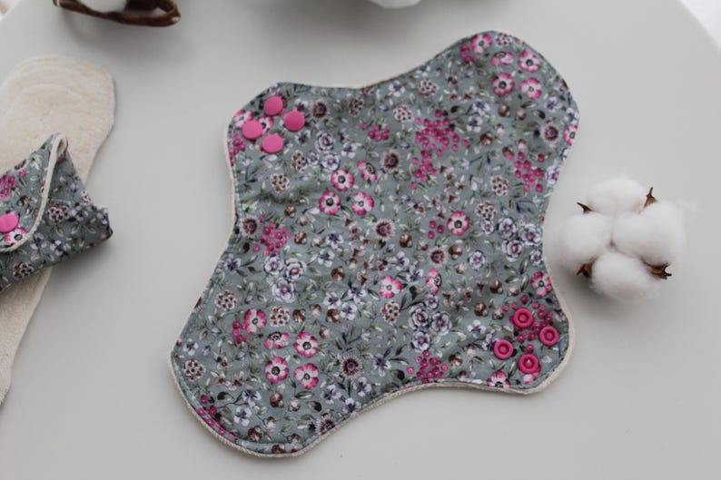 May include: Reusable cloth menstrual pads with a floral pattern in shades of gray, pink, and white. The pads have pink snap closures. A cotton ball and other pads are also visible.