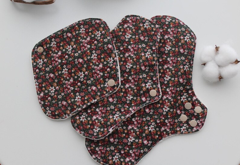 May include: Reusable cloth menstrual pads in a floral pattern. The pads are dark brown with a small white and pink floral print. The pads have snap closures and are in various shapes and sizes.