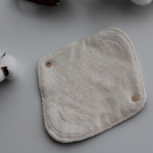 May include: A square, off-white reusable cloth pad with rounded corners and two snaps. A cotton boll is visible in the upper left corner. The pad is made of soft, absorbent material, ideal for personal hygiene.