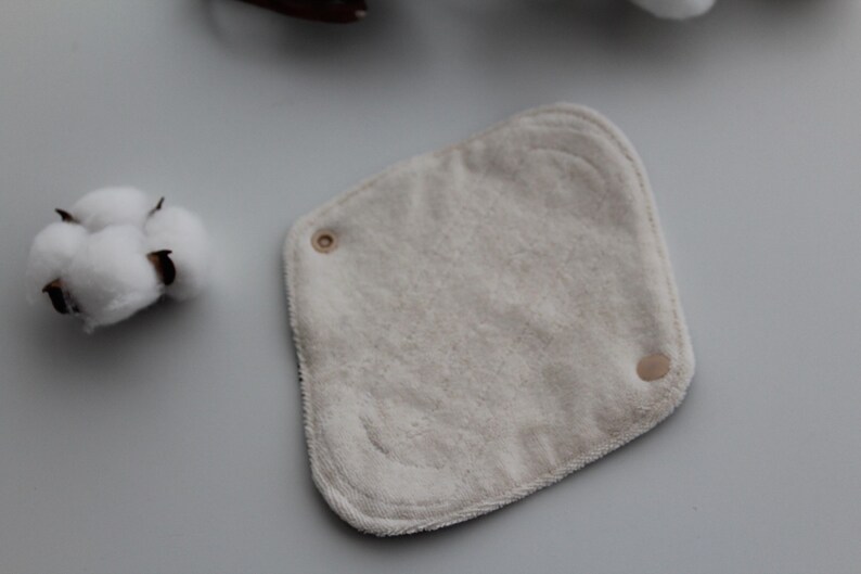 May include: A square, off-white reusable cloth pad with rounded corners and two snaps. A cotton boll is visible in the upper left corner. The pad is made of soft, absorbent material, ideal for personal hygiene.