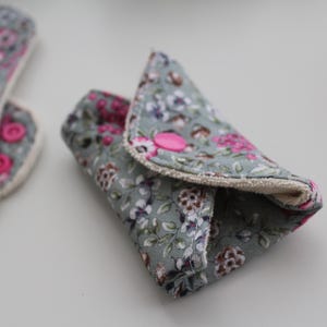 May include: Reusable cloth menstrual pads with a floral pattern in shades of gray, pink, and white. One pad is folded and secured with a pink snap closure. Another pad is visible in the background.