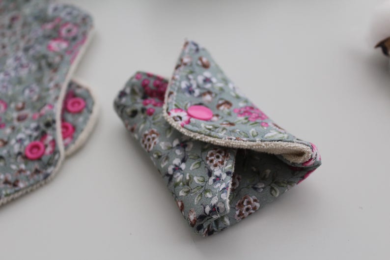 May include: Reusable cloth menstrual pads with a floral pattern in shades of gray, pink, and white. One pad is folded and secured with a pink snap closure. Another pad is visible in the background.