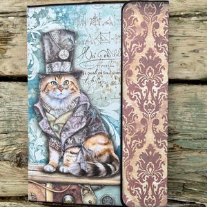 May include: A decorative file folder featuring a whimsical illustration of a cat dressed in Victorian attire, including a top hat and coat. The folder has a blue background with script and geometric designs, and a patterned spine in shades of brown and beige.
