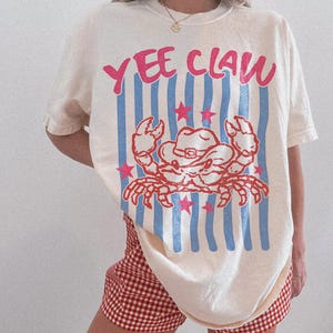Könnte beinhalten: Cremefarbenes T-Shirt mit dem Text "YEE CLAW" in Pink, über einer roten Krabbe mit Cowboyhut. Das Design umfasst blaue vertikale Streifen und rosa Sterne. Das Shirt wird mit rot-weiß karierten Shorts kombiniert.