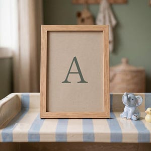 May include: A wooden framed print displaying the letter "A" in green. The frame rests on a striped surface, alongside a small blue elephant and a yellow duck figurine. The setting suggests a nursery or children's room.