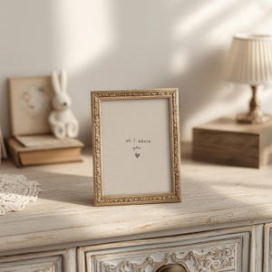 May include: A gold-coloured rectangular picture frame with decorative detailing. The frame contains a beige insert with the handwritten text "oh I adore you" and a small heart. The frame is on a white dresser.