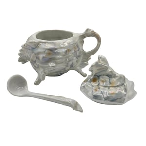 May include: A ceramic sugar bowl with a lid and a matching spoon, featuring a glossy, iridescent finish with a subtle floral pattern in shades of white, gray, and soft orange. The bowl stands on three small legs and has a curved handle.