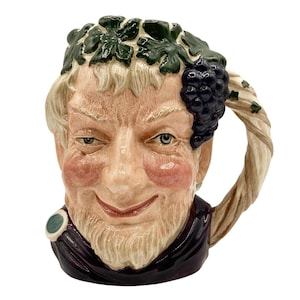 May include: A ceramic character mug shaped like a person's head with a mischievous expression, featuring a crown of dark green grape leaves and a cluster of purple grapes. The handle is designed to look like a twisted vine, and the base is dark purple.