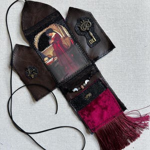 May include: A brown faux leather hanging ornament featuring a central rectangular print of a figure in a red robe, accented with a brass-coloured key charm, a star emblem, a moon phase bead strand, a black glitter panel, and a deep red velvet tassel.