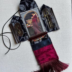 May include: A fabric wall hanging featuring a central image of a person in a red dress, flanked by two side panels with black floral fabric, a bronze moon charm, and a bronze key charm with the word Love. It includes a deep red velvet base with fringe.