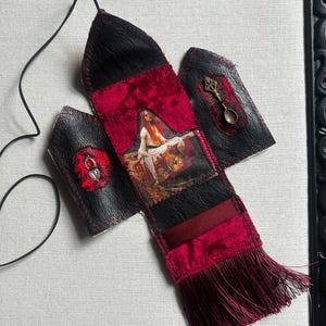 May include: A black leather pouch with a red velvet centre panel featuring a printed image of a person in white clothing. The pouch includes a red tassel, a small bronze-coloured spoon charm, a red gemstone pendant, and a thin black cord for hanging.