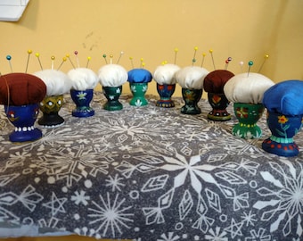 May include: A row of 10 colorful pincushions, each with a white fabric top and a painted egg-shaped base. The bases are painted in various colors and patterns, including blue, green, red, and white.