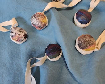 May include: Five colorful fabric balls with abstract patterns are strung together on a white ribbon. The balls are tied together with a knot and are hanging on a blue fabric background.