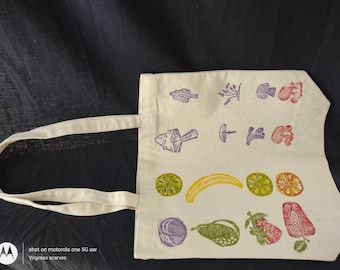 May include: A white canvas tote bag with a variety of colorful hand-stamped images of fruits, vegetables, and mushrooms. The bag has a long handle and is made of natural fabric.