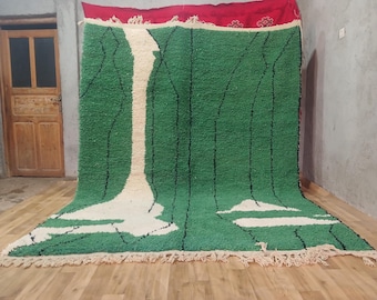 May include: A large, green and white rug with a geometric design. The rug features a central white pattern with black lines and a green background. The top edge is red with a decorative border. The rug is displayed on a wooden floor.
