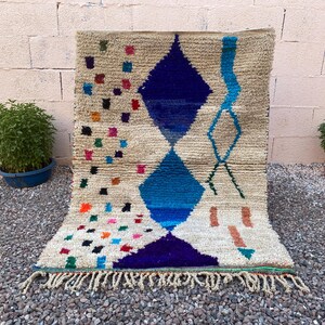 May include: A beige Moroccan rug with a geometric design. The rug features a large blue diamond pattern, with smaller colorful squares and fringe along the bottom. The rug is placed on a gravel surface.