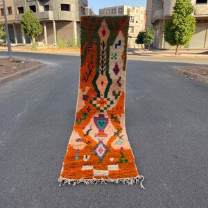 May include: A long, rectangular runner rug featuring a vibrant orange base with a cream-colored center panel. The rug is decorated with intricate geometric patterns in shades of dark green, purple, pink, and blue, finished with white fringe.