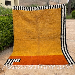 May include: A rectangular, handmade rug with a textured, golden-yellow field. The bottom section is orange, and the top and sides have black and white striped borders. White fringe accents the bottom edge. The rug is approximately 180 cm wide and 240 cm long.