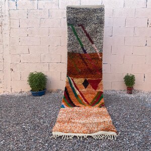 May include: A rectangular, vertically oriented rug with a geometric design. The rug features a mix of colors, including orange, brown, green, and blue. The top portion is a speckled gray, transitioning to orange and brown. The bottom has a fringed edge.