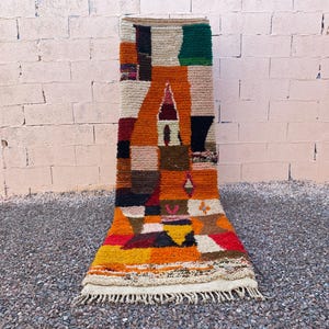 Könnte beinhalten: Rechteckiger Teppich mit geometrischem Muster in Orange, Rot, Grün und Braun. Der Teppich hat einen Fransenrand und wird vor einer Backsteinmauer präsentiert. Das Design ist bunt und abstrakt.